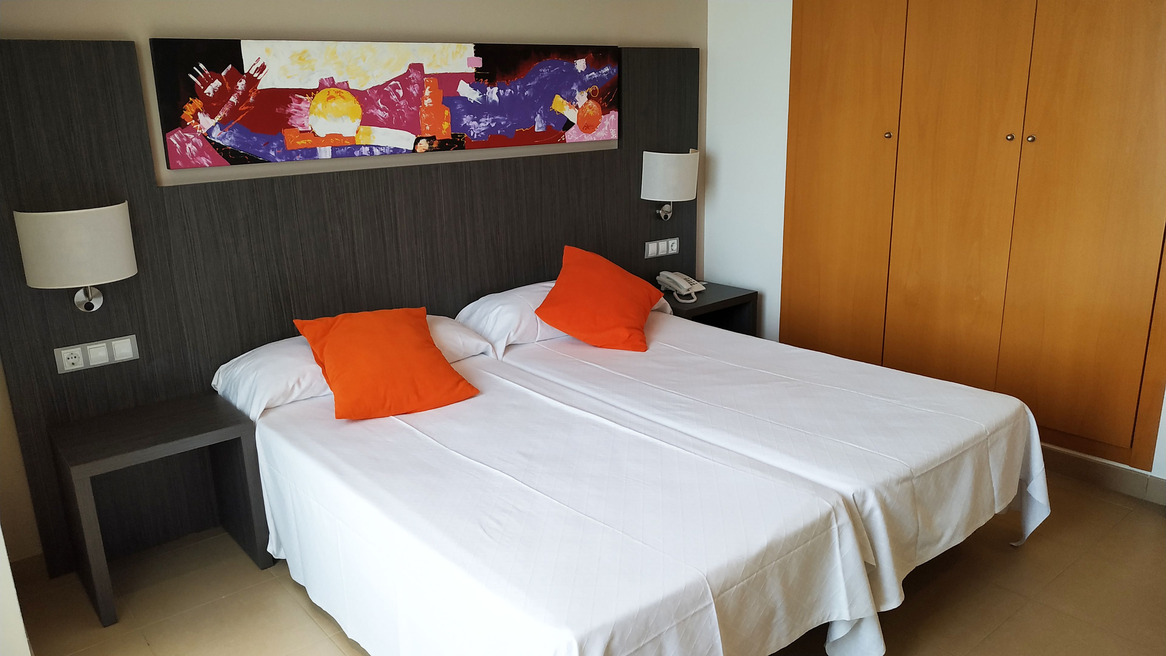 A hotel room with a double bed, two nightstands, a telephone, a wall-mounted lamp, and a wooden wardrobe.
