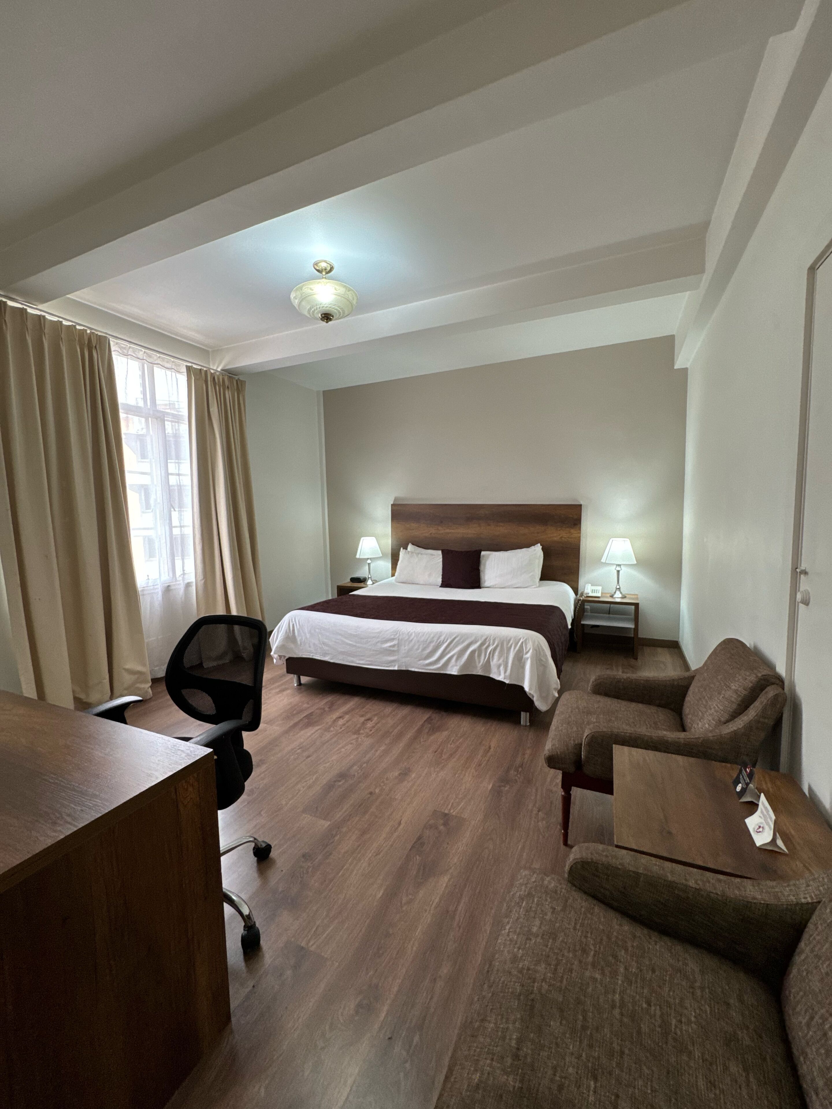Una camera d'albergo con un letto, due comodini con lampade, una scrivania in legno, una sedia e un'ampia finestra con tende.