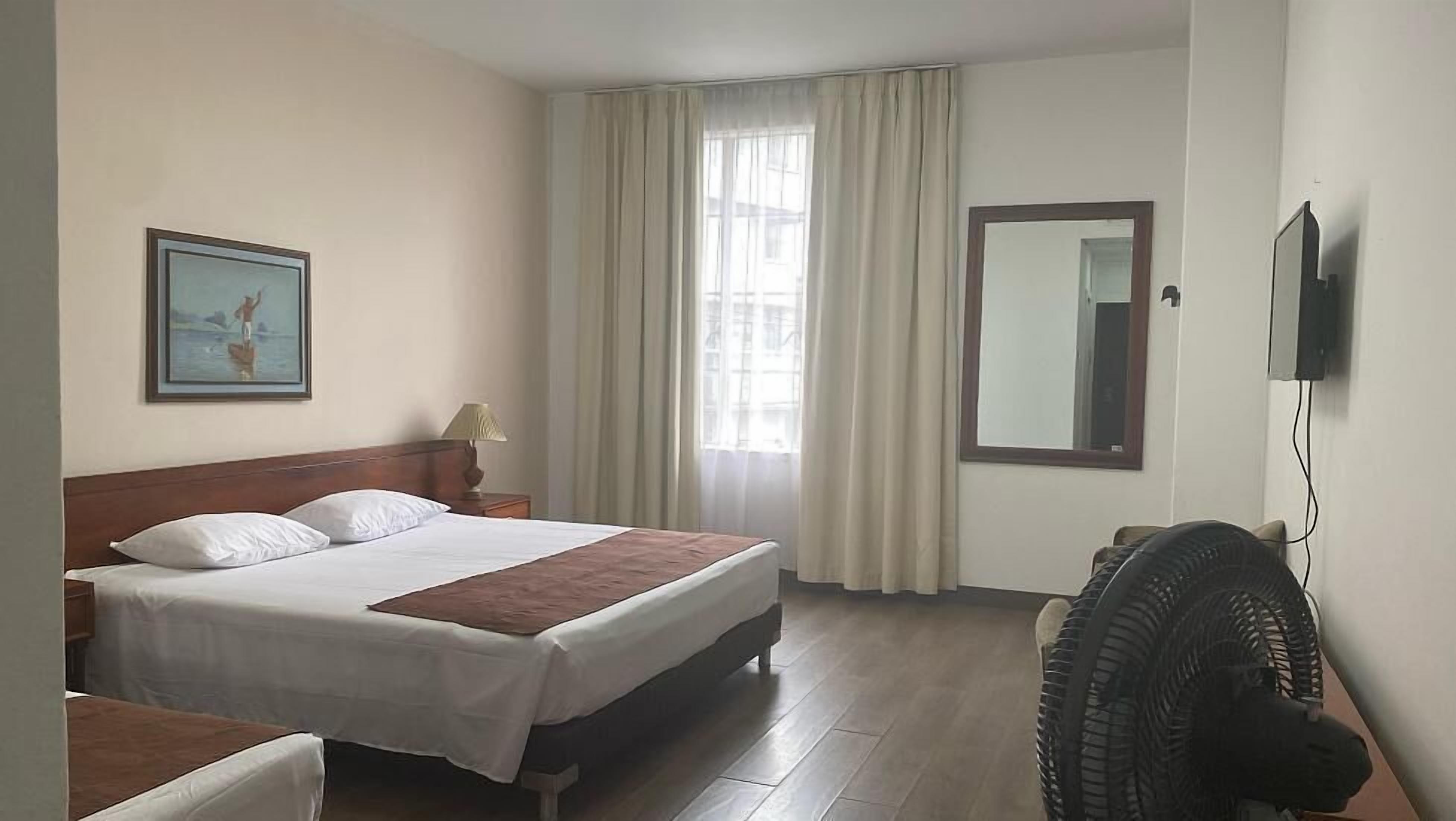 A hotel room with a bed, a bedside table, a lamp, a mirror, a window with curtains, and a wall-mounted TV.
