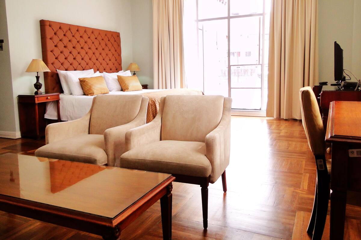A hotel room with a large bed, two armchairs, a wooden coffee table, a desk with a computer, and a window with curtains.