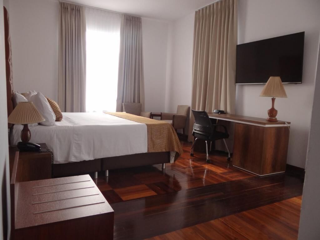 A hotel room with a bed, a desk, a chair, a TV, and a lamp.