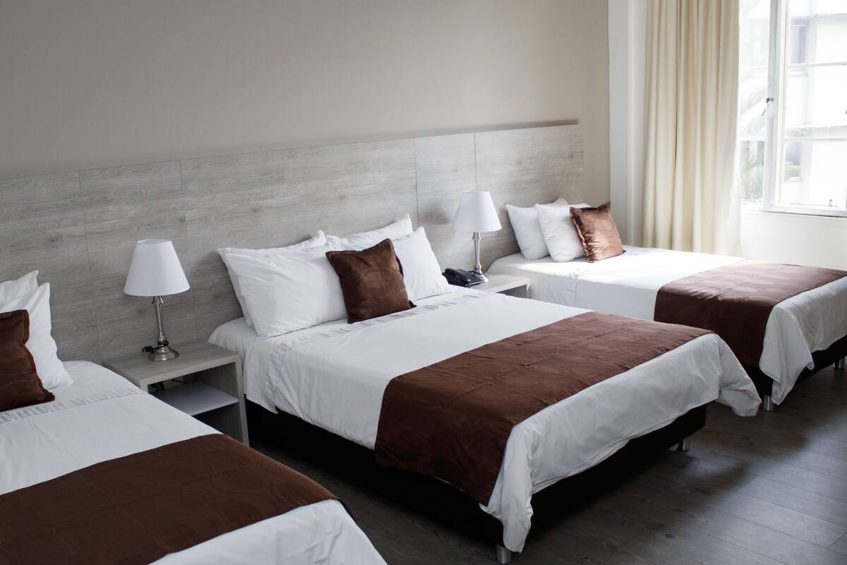 A hotel room with three beds, each with white bedding and brown throw pillows. There is a nightstand with a lamp and a phone on each bed. The room has a wooden headboard and a window with curtains.