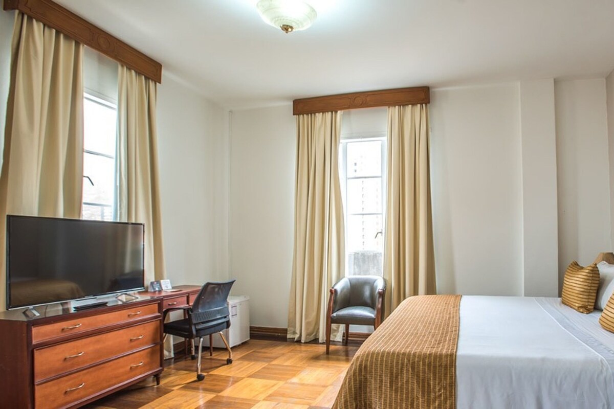 Une chambre d’hôtel avec un grand lit, une télévision, un bureau avec une chaise et une fenêtre avec des rideaux.