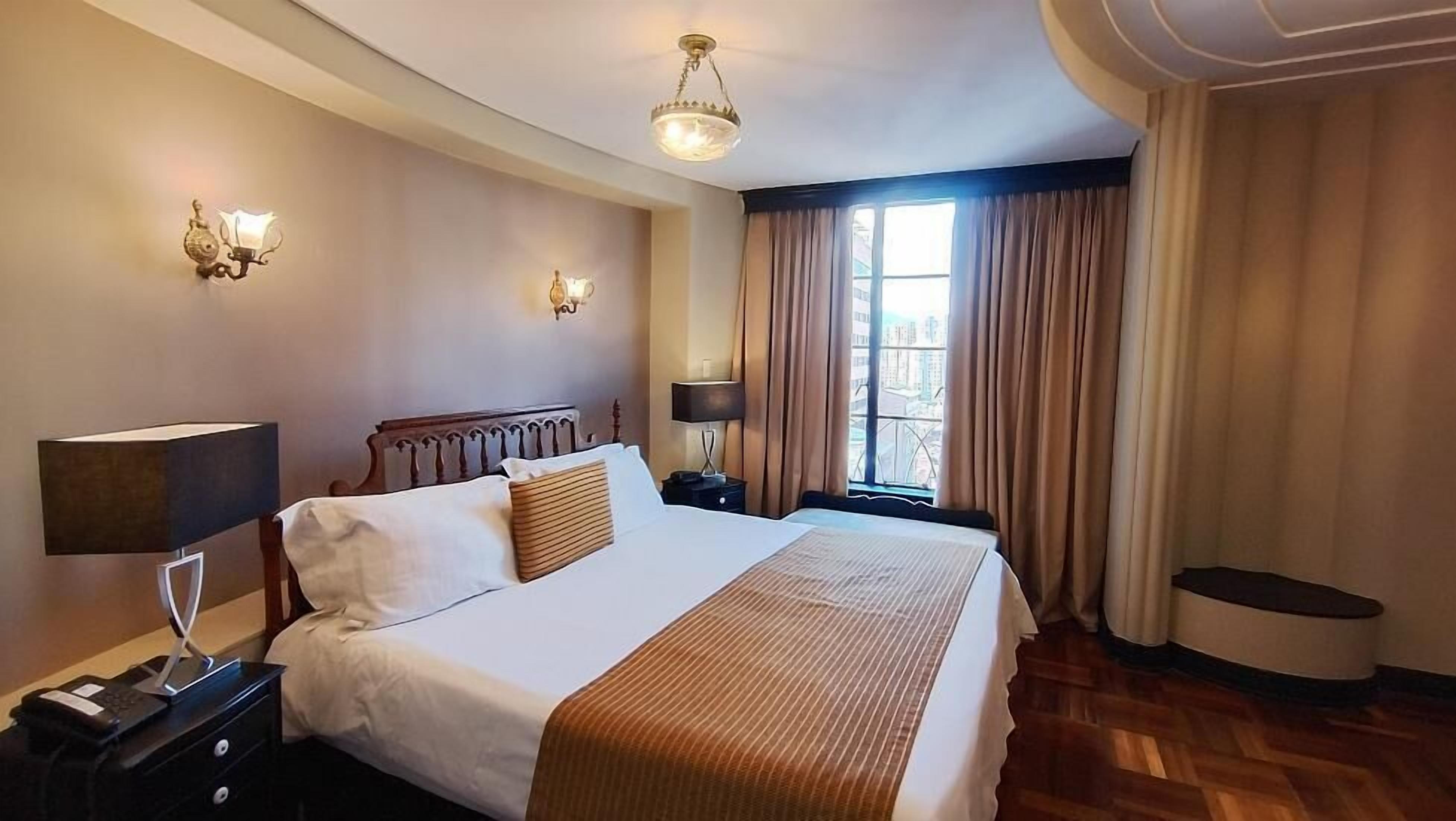 Habitación de hotel con una cama grande, lámparas de noche, mesita de noche, una ventana con cortinas y un candelabro.