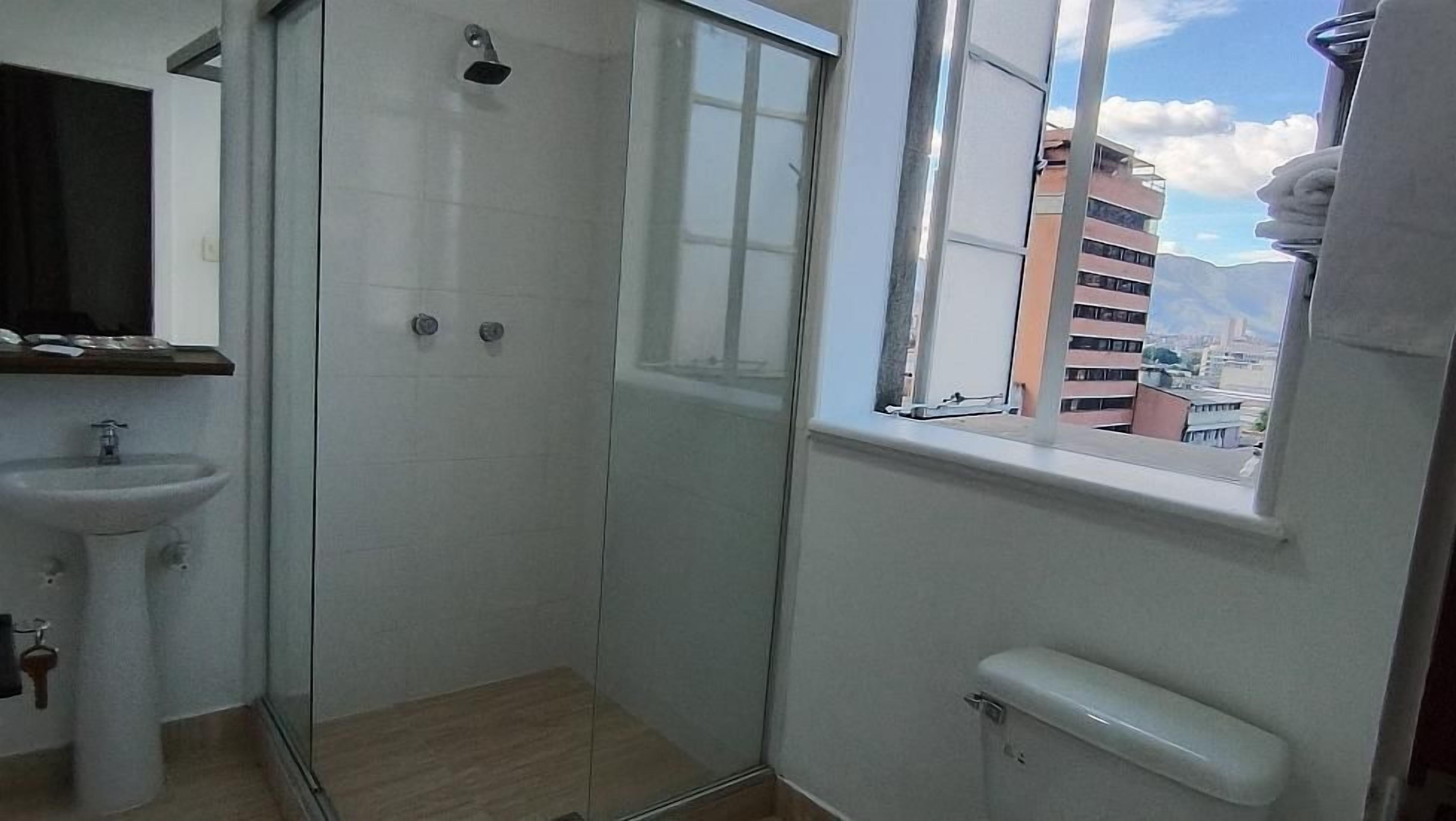 Une salle de bain avec une douche à parois de verre, un lavabo et une fenêtre donnant sur des immeubles.