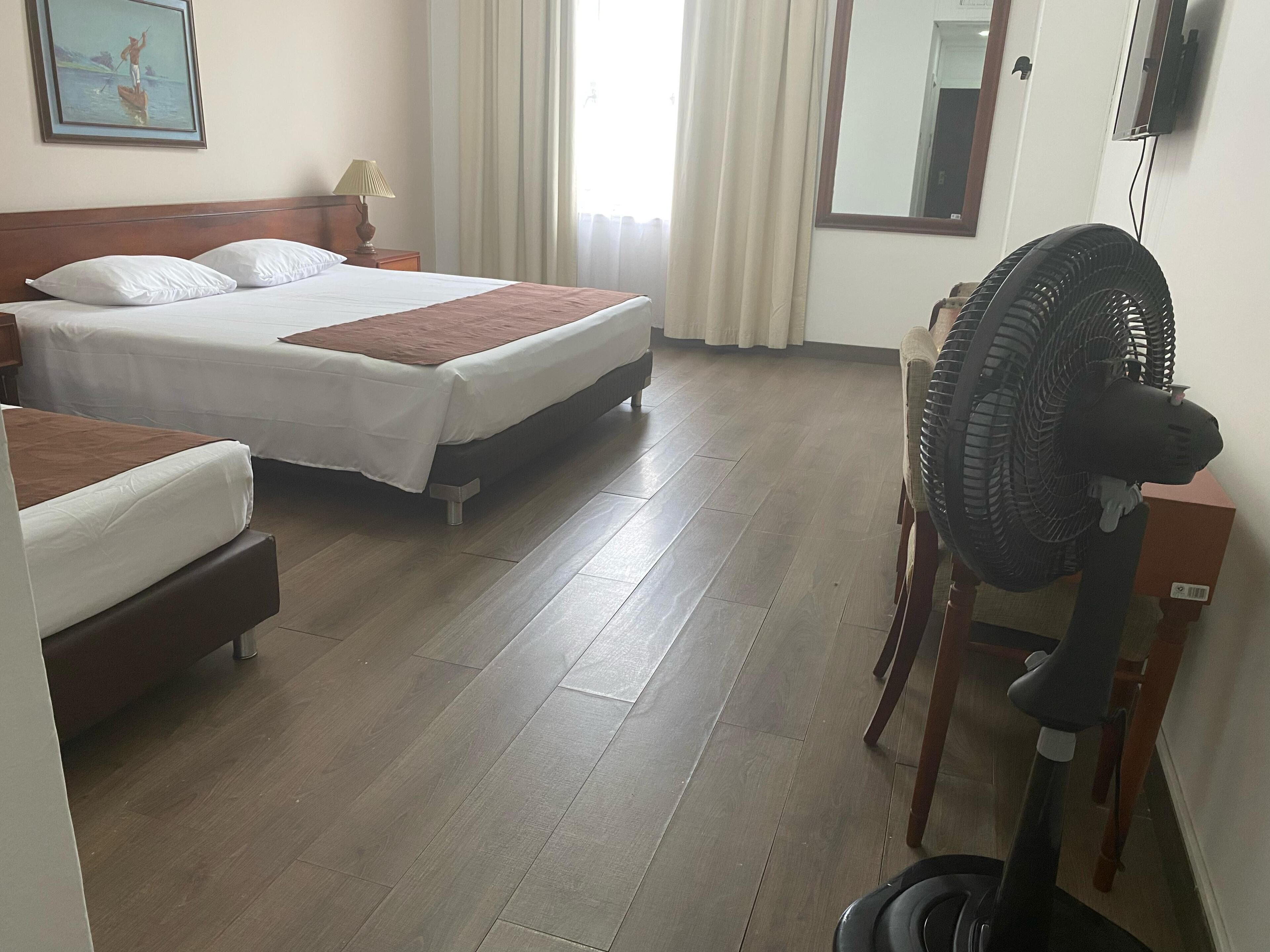 Hotellihuone, jossa on kaksi sänkyä, lattiatuuletin ja puulattia.