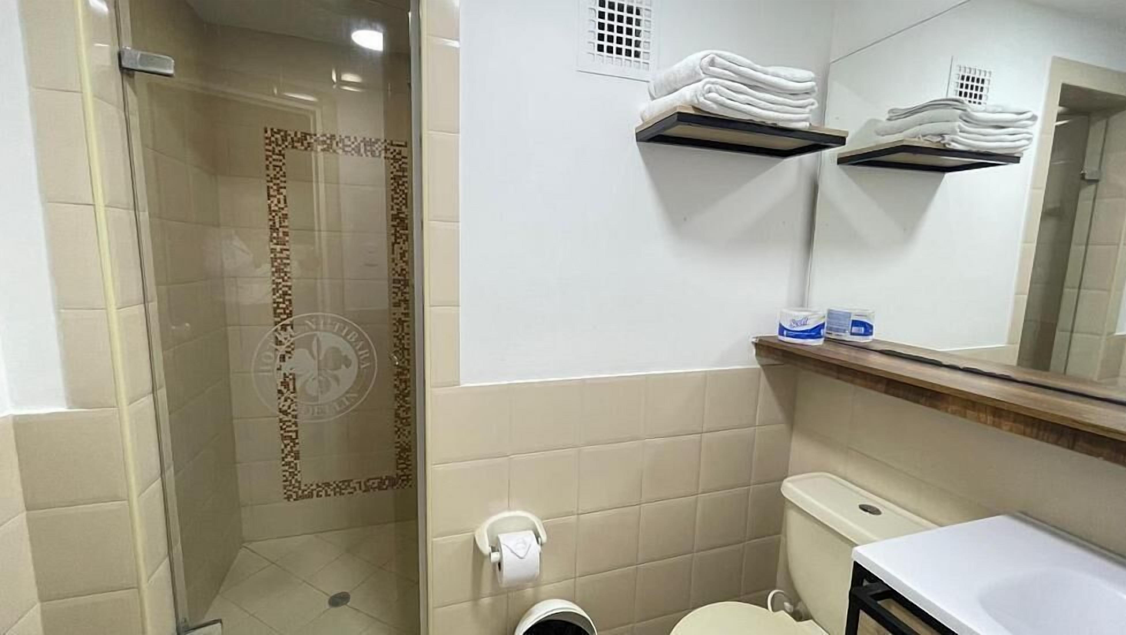 Une salle de bain avec une douche à parois de verre, des toilettes, un lavabo et des étagères où sont pliées des serviettes.