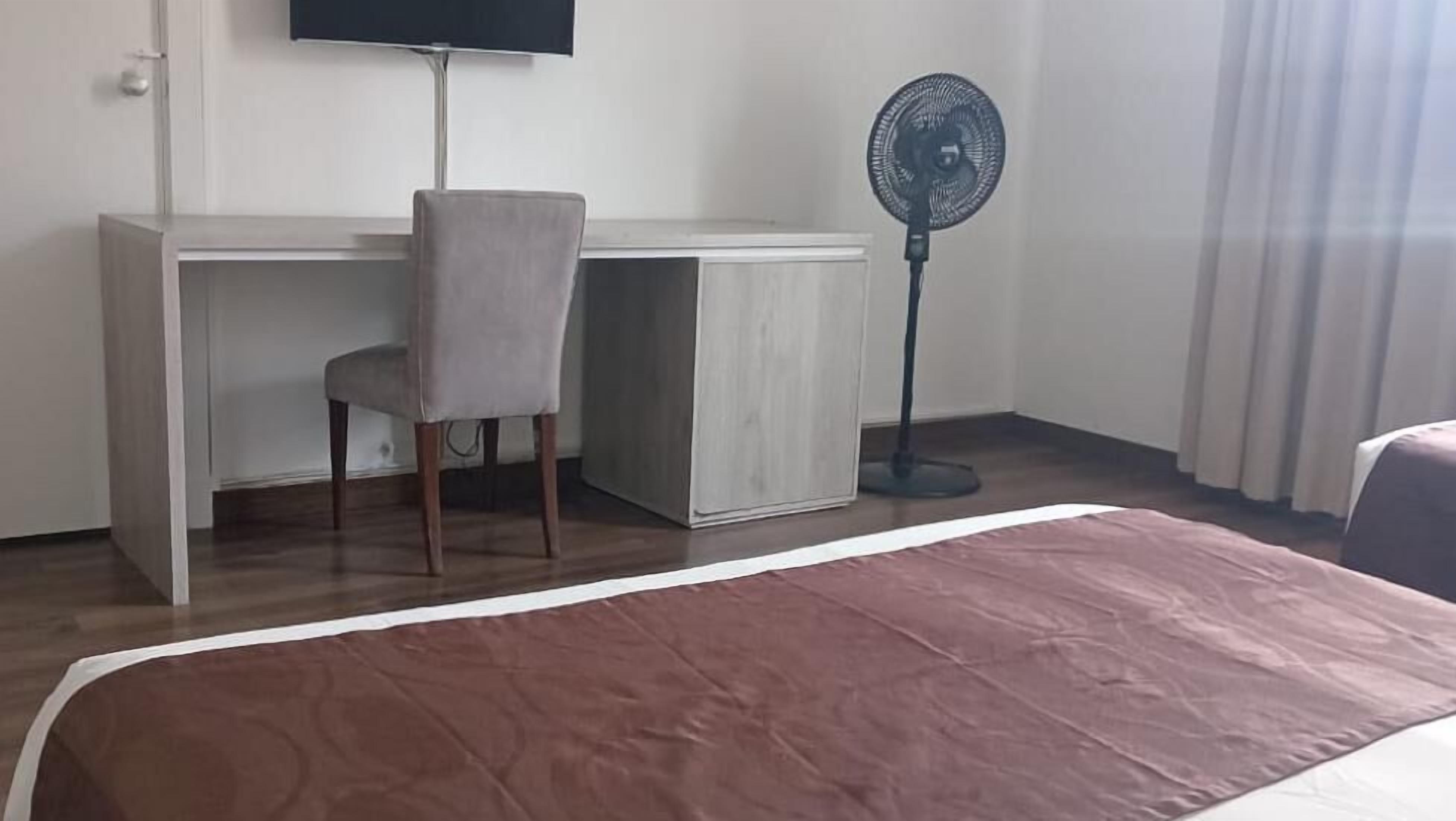 Ein Zimmer mit einem Bett, einem Schreibtisch, einem Stuhl und einem Standventilator.
