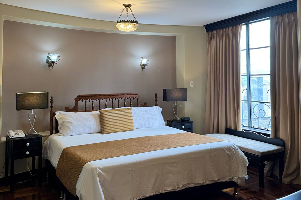 A hotel room with a large bed, two bedside tables, a chandelier, and a window with curtains.