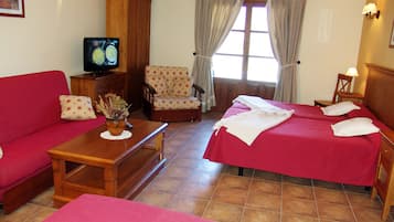 Hotelkamer met een rode bank, een houten bijzettafeltje met een vaas, een fauteuil met bloemenprint, twee eenpersoonsbedden met witte lakens, een houten garderobe, een televisie op een houten dressoir en een raam met gordijnen.