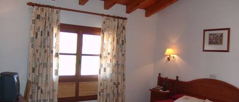 A bedroom with a wooden ceiling, a bed with red bedding, a nightstand with a lamp, and a window with patterned curtains.