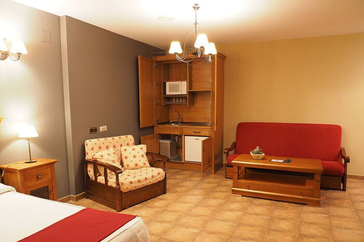 Habitación compacta con cama, sofá rojo, silla de madera, pequeña mesa, kitchenette y candelabro.