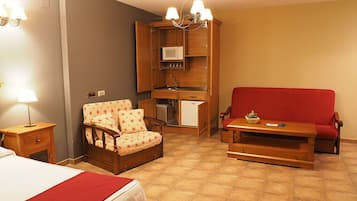 A compact room with a bed, a red sofa, a wooden chair, a small table, a kitchenette, and a chandelier.