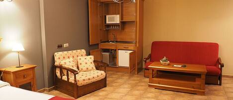 A compact room with a bed, a red sofa, a wooden chair, a small table, a kitchenette, and a chandelier.