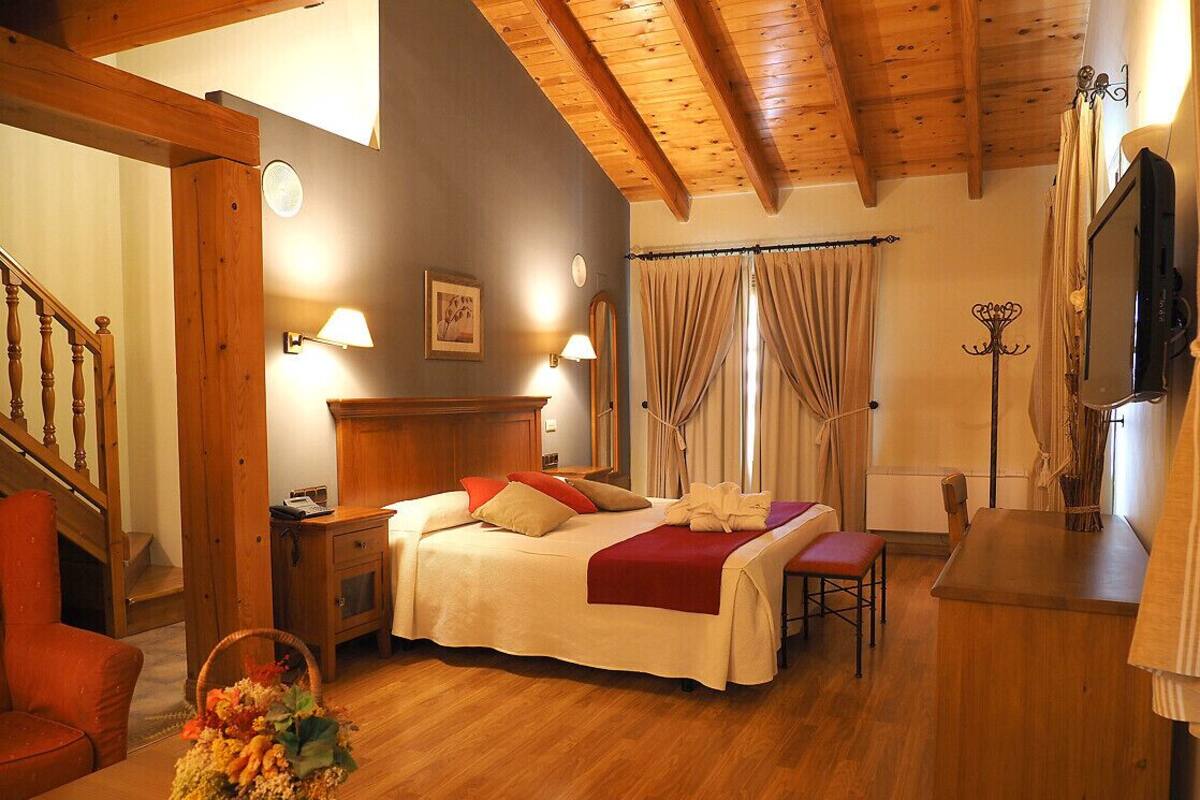 Habitación de hotel con techo de madera, cama con manta roja, mesita de noche con lámpara, una silla y un escritorio de madera con televisor.