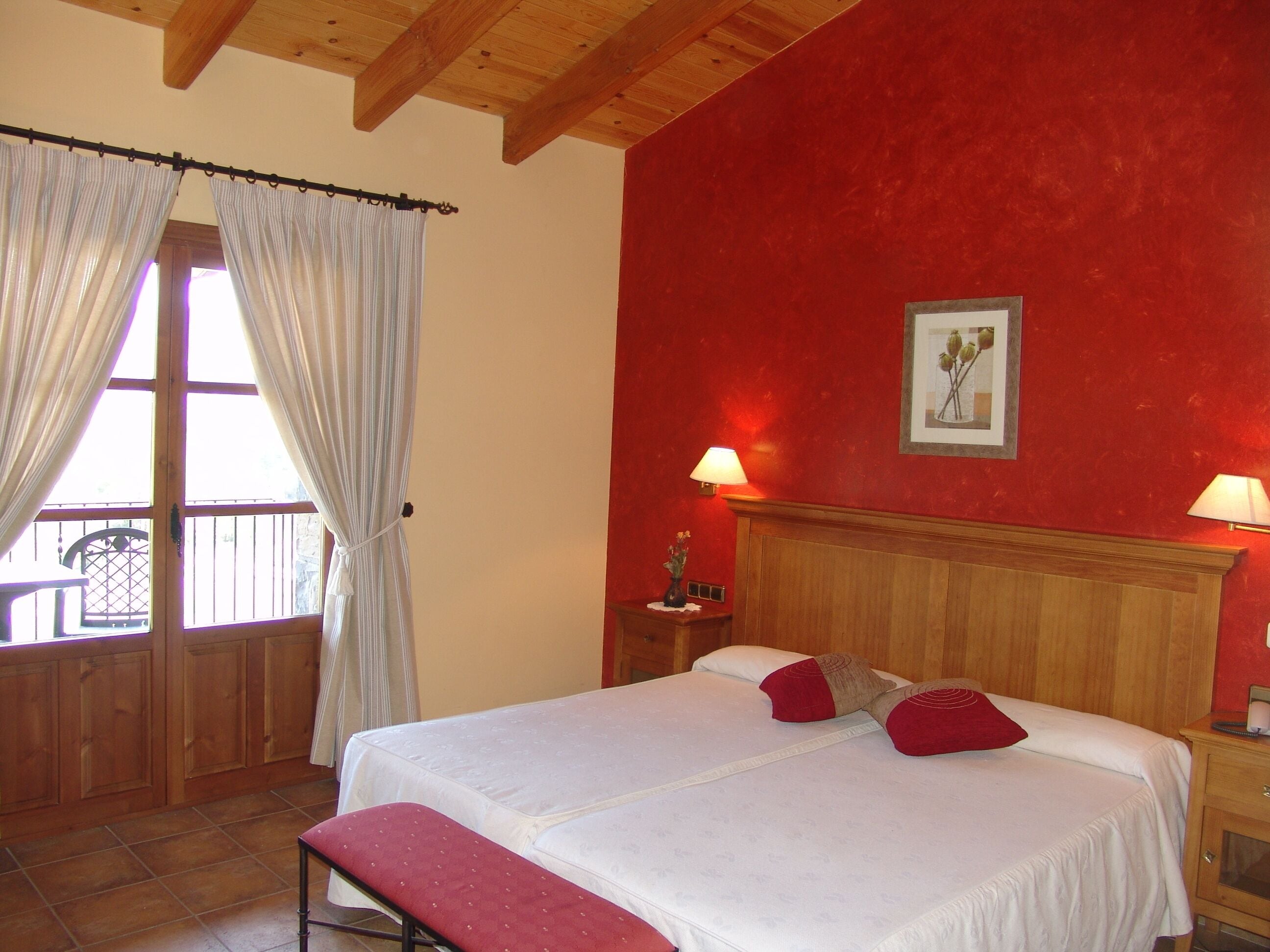 Un dormitorio con cabecera de madera, cama con ropa blanca y almohadas rojas, una banca al pie de la cama, dos lámparas de noche y una ventana con cortinas.