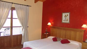 Un dormitorio con cabecera de madera, cama con ropa blanca y almohadas rojas, una banca al pie de la cama, dos lámparas de noche y una ventana con cortinas.