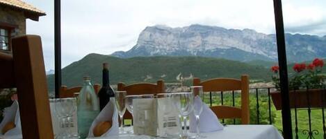 Mesa de comedor servida con vasos, platos y una botella, con vistas a un paisaje montañoso.