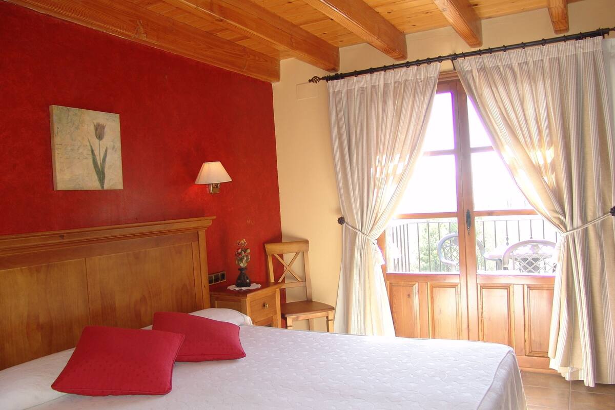 Un dormitorio con cabecera de madera, cama con ropa de cama blanca y dos almohadas rojas, mesita de noche con lámpara, un cuadro de una flor en la pared y un balcón con silla y barandilla.