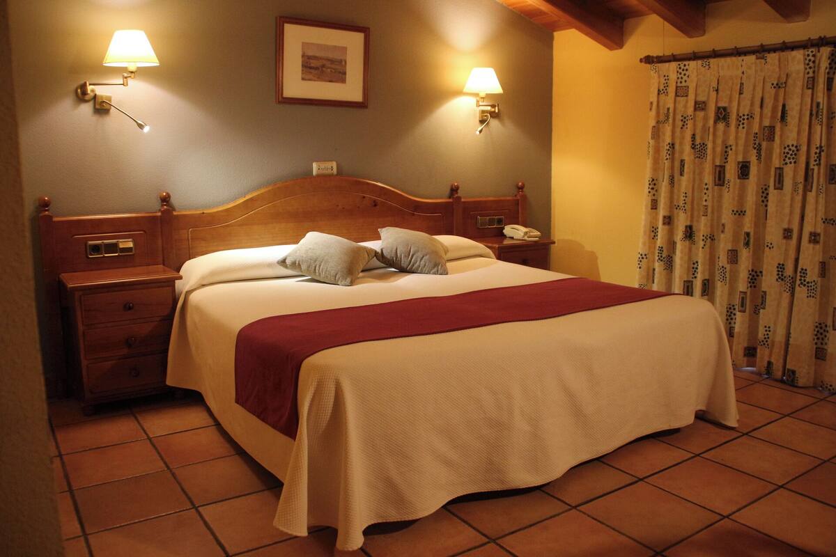 Una cama bien hecha con una colcha roja, dos lámparas de noche, una mesita de noche de madera con un teléfono y un cuadro en la pared.