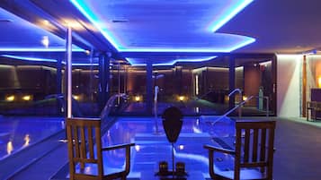 Un área de piscina interior moderna con paredes de vidrio, sillas de madera e iluminación ambiental azul.
