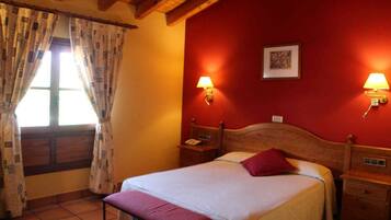Un dormitorio con cabecera de madera, una cama individual con almohada roja, dos mesitas de noche con lámparas, una ventana con cortinas estampadas y una pared roja con un cuadro enmarcado.