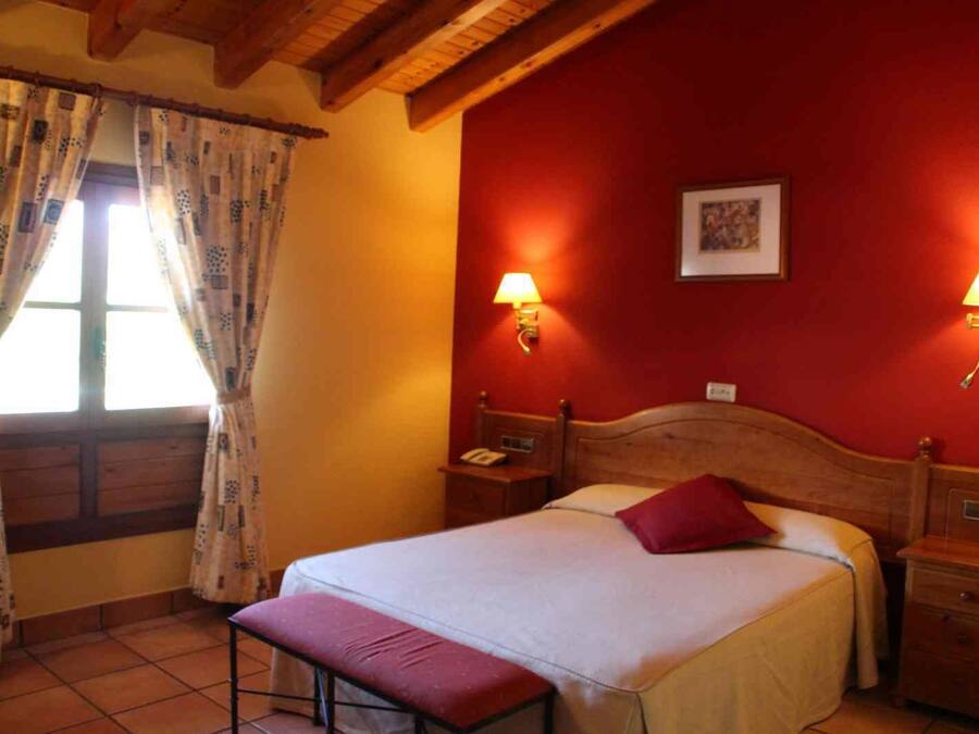 Un dormitorio con cabecera de madera, una cama individual con almohada roja, dos mesitas de noche con lámparas, una ventana con cortinas estampadas y una pared roja con un cuadro enmarcado.