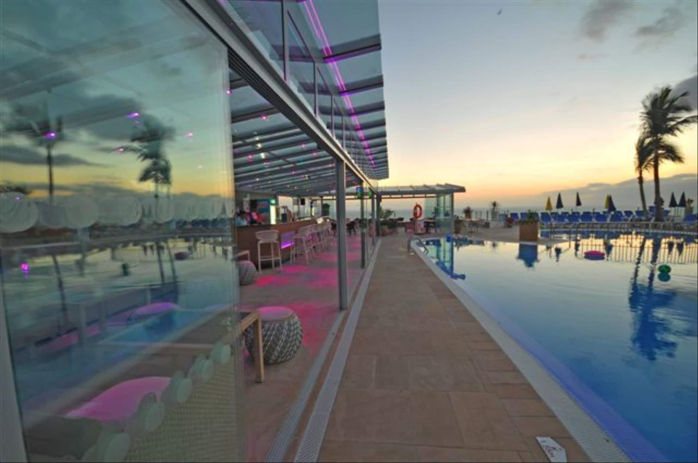 Zona junto a la piscina con cerramiento de vidrio, sillas de descanso y vista al atardecer.