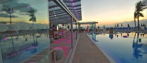 Zona junto a la piscina con cerramiento de vidrio, sillas de descanso y vista al atardecer.
