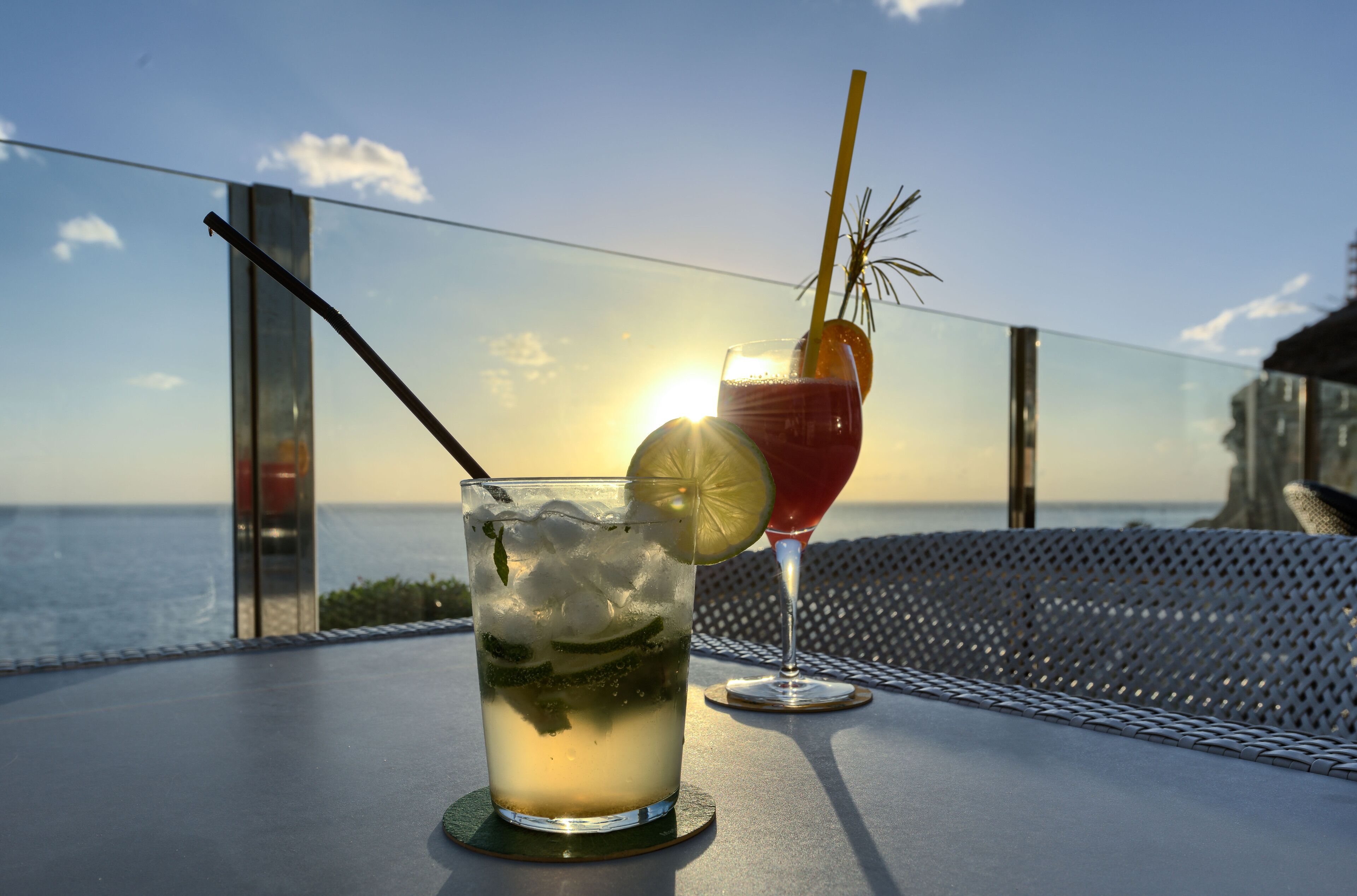Two drinks on a table with a view of the sea and a building in the background.