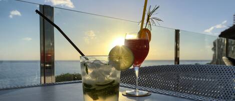 Two drinks on a table with a view of the sea and a building in the background.
