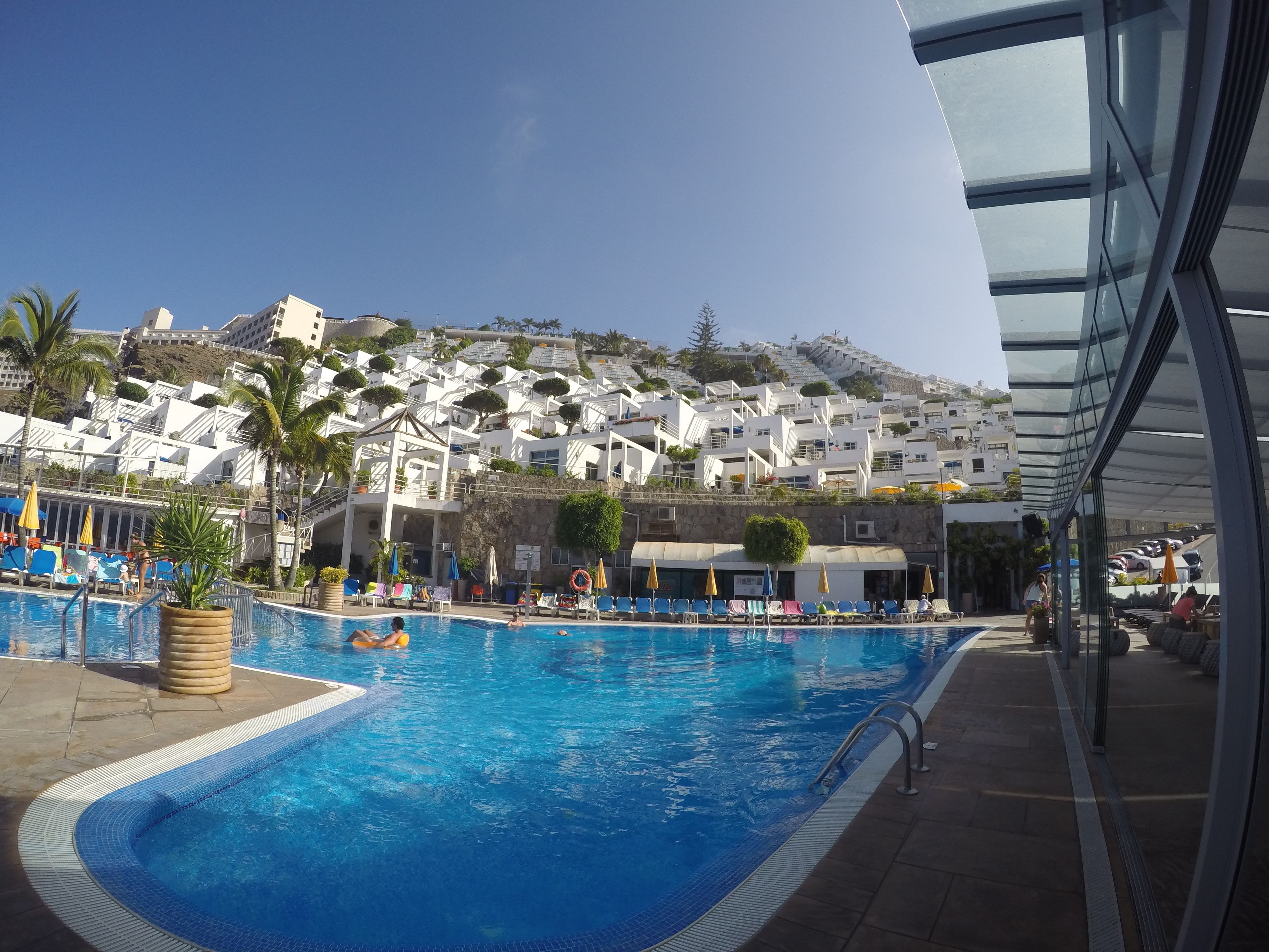 Une piscine entourée de chaises longues et de parasols, bordée de bâtiments blancs dotés de balcons, avec une colline en arrière-plan.