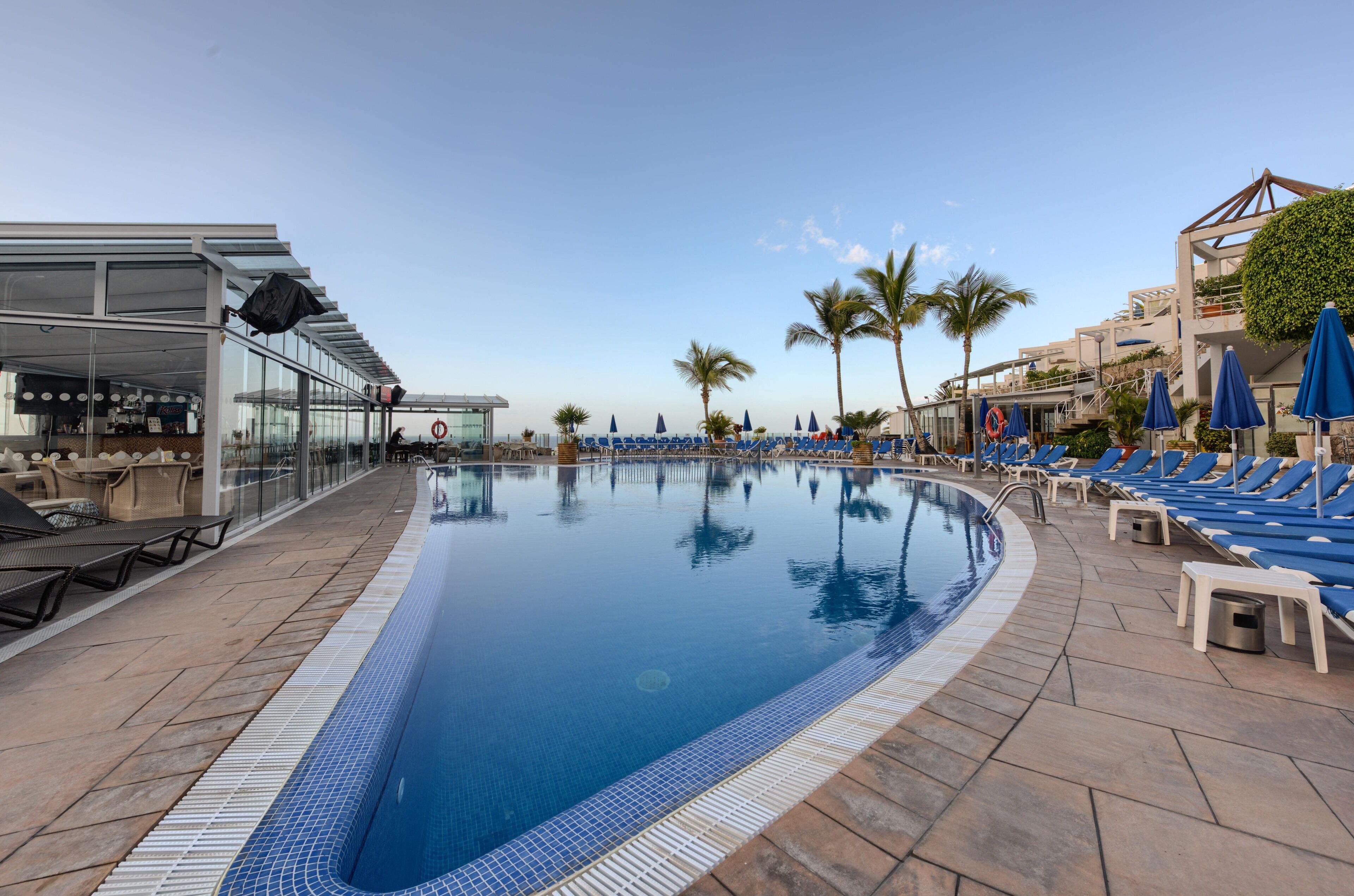 Ein Swimmingpool mit Liegestühlen und Sonnenschirmen, umgeben von Palmen und einem modernen Gebäude.