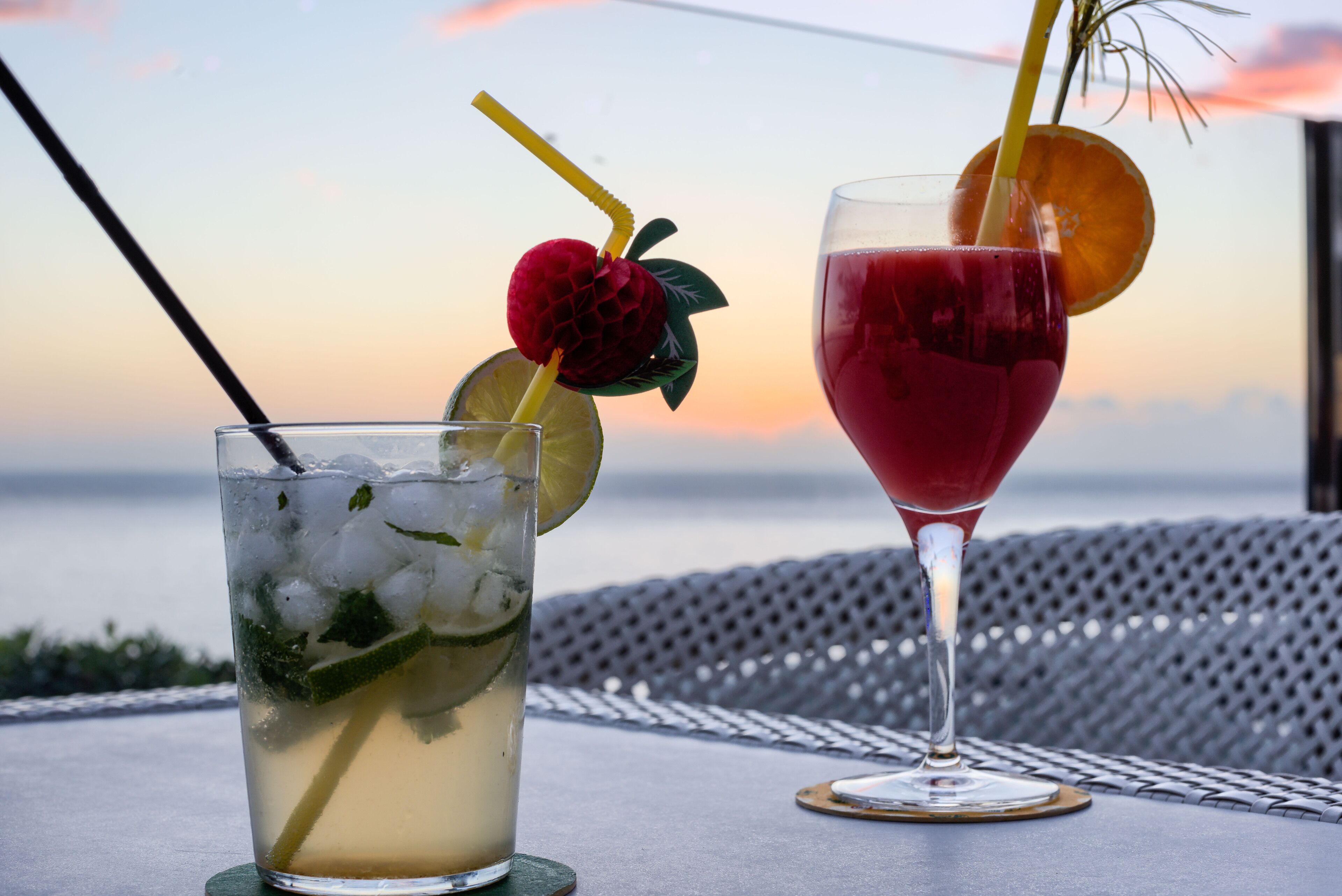 Two drinks on a table with a view of the sea and a sunset in the background.