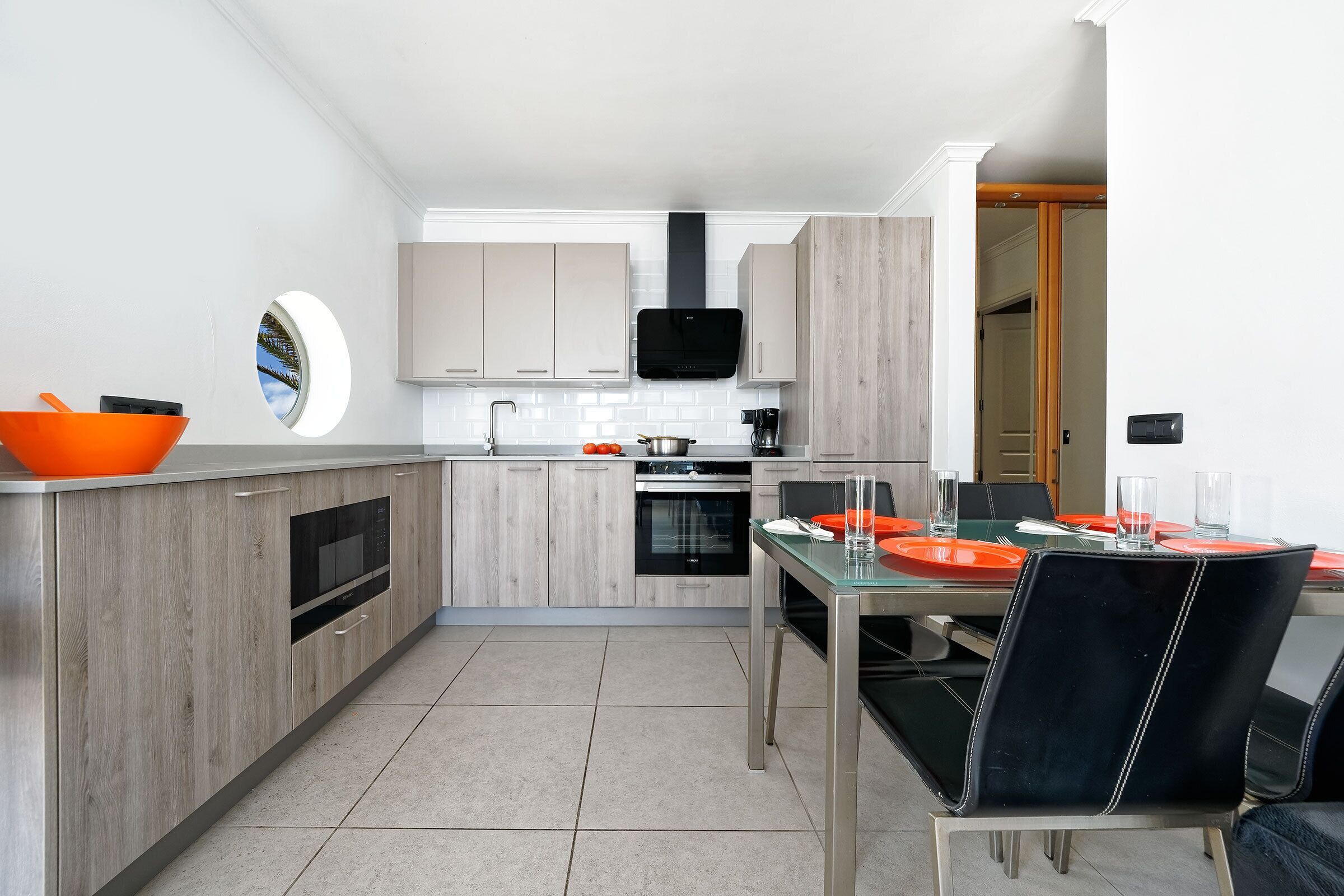 Une cuisine moderne avec des meubles en bois, un four encastré et un coin repas comprenant une table et des chaises en verre.