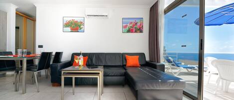 A modern living room with a sofa, a coffee table, and a dining area with chairs. There are two paintings on the wall and a large window with a view of the ocean.