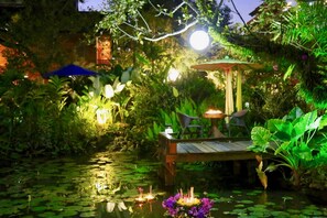 Un jardín tranquilo con un muelle de madera sobre un estanque, rodeado de abundante vegetación y un pabellón con mesa y sillas.