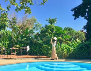 Un área junto a la piscina con una piscina azul cristalina, rodeada de exuberante vegetación y diversas plantas.