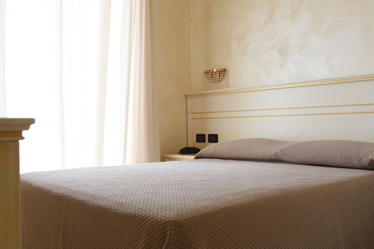Un lit bien fait, recouvert d’une couverture à motifs, une table de chevet avec une lampe et un luminaire fixé au mur.