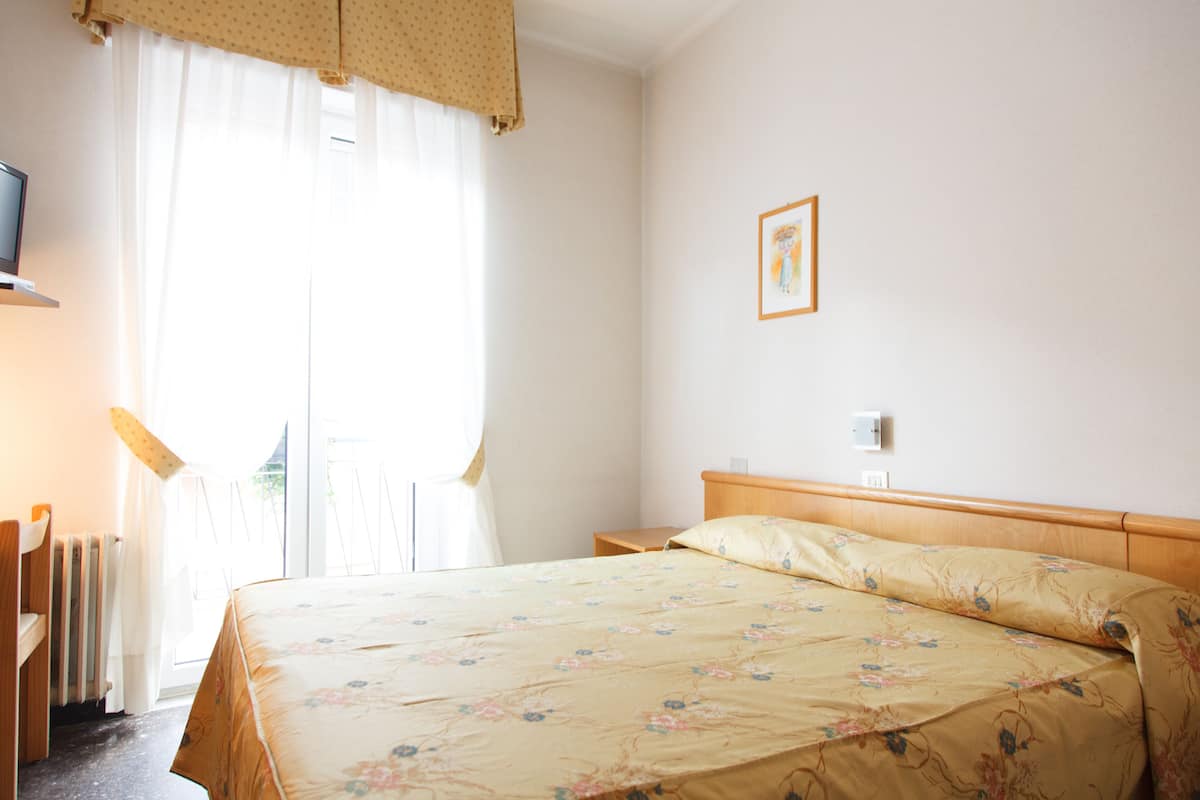 Une chambre d’hôtel avec un lit, une table de chevet, une lampe, un radiateur et une fenêtre avec des rideaux.