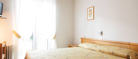 Quarto de hotel com uma cama, mesa de cabeceira, abajur, radiador e janela com cortinas.