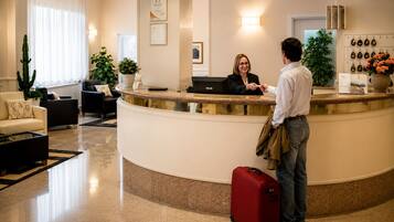 Zona de recepción de un hotel con un mostrador curvo, una recepcionista, un huésped con una maleta roja y una pared con diplomas enmarcados.