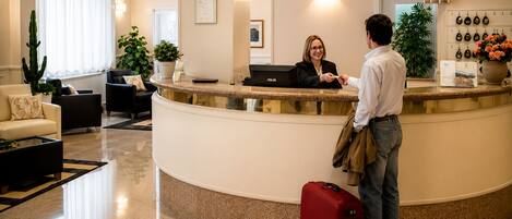 Zona de recepción de un hotel con un mostrador curvo, una recepcionista, un huésped con una maleta roja y una pared con diplomas enmarcados.