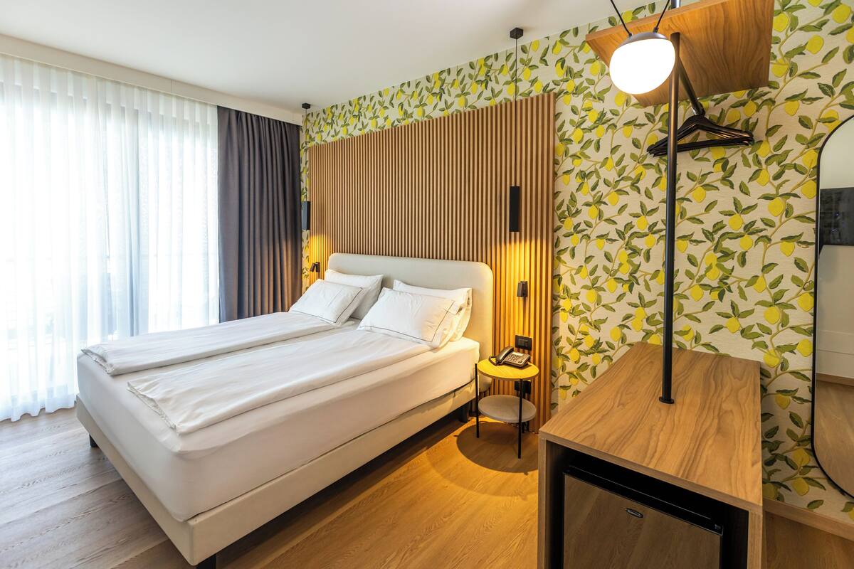 Une chambre d’hôtel moderne avec un grand lit, une table de chevet avec une lampe et un mur orné d’un motif de feuilles.