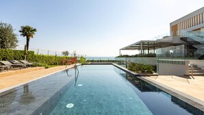 Una piscina infinita moderna con agua cristalina, rodeada por una terraza de madera y barandillas de vidrio, con vista al litoral.