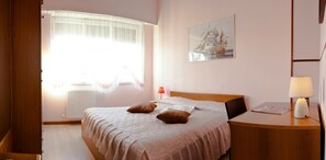 Un dormitorio ordenado con cama, mesita de noche con lámpara, ventana con cortinas y un cuadro colgado en la pared.