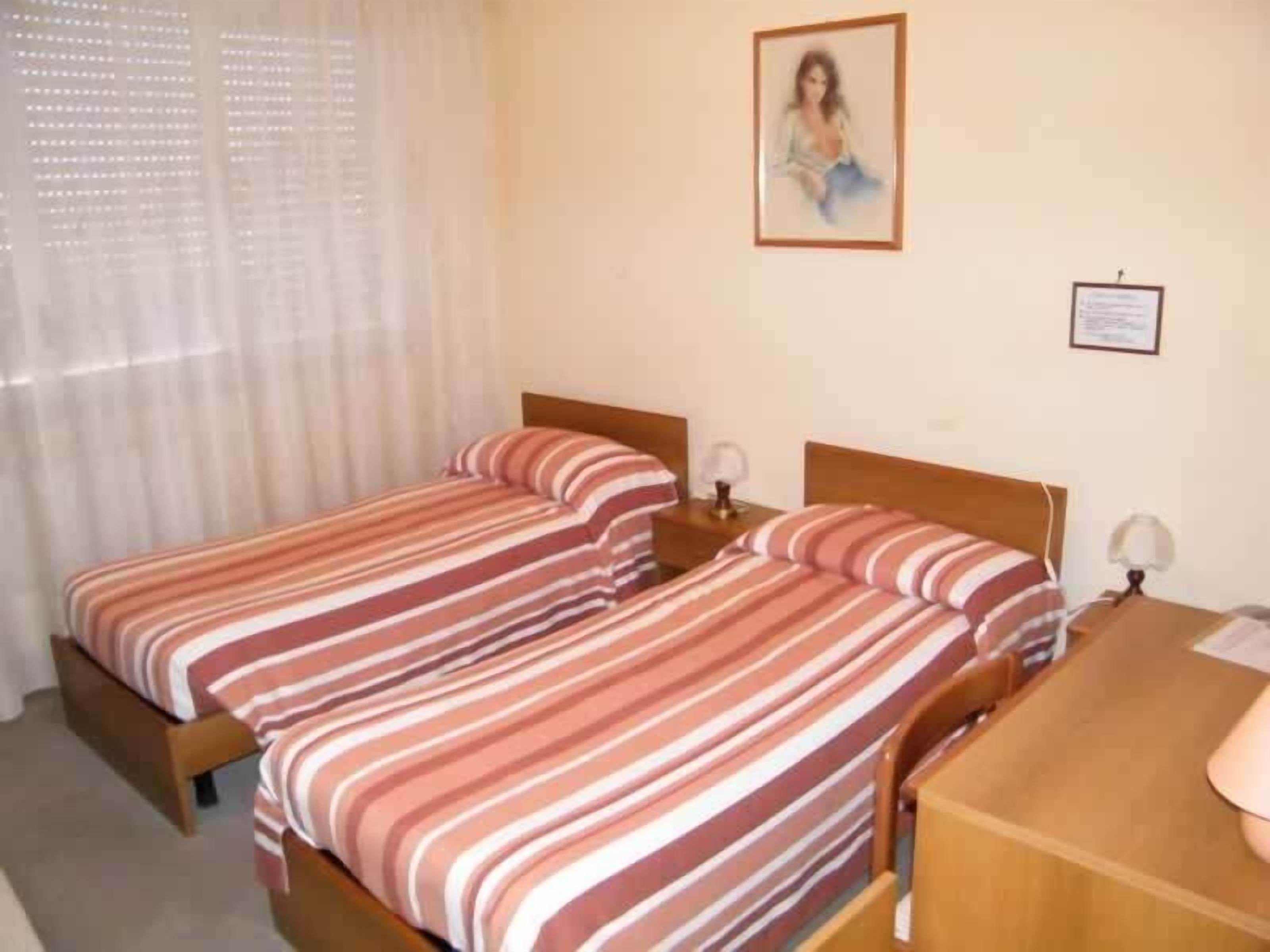 A hotel room with two beds, a wooden desk, and a window with sheer curtains.
