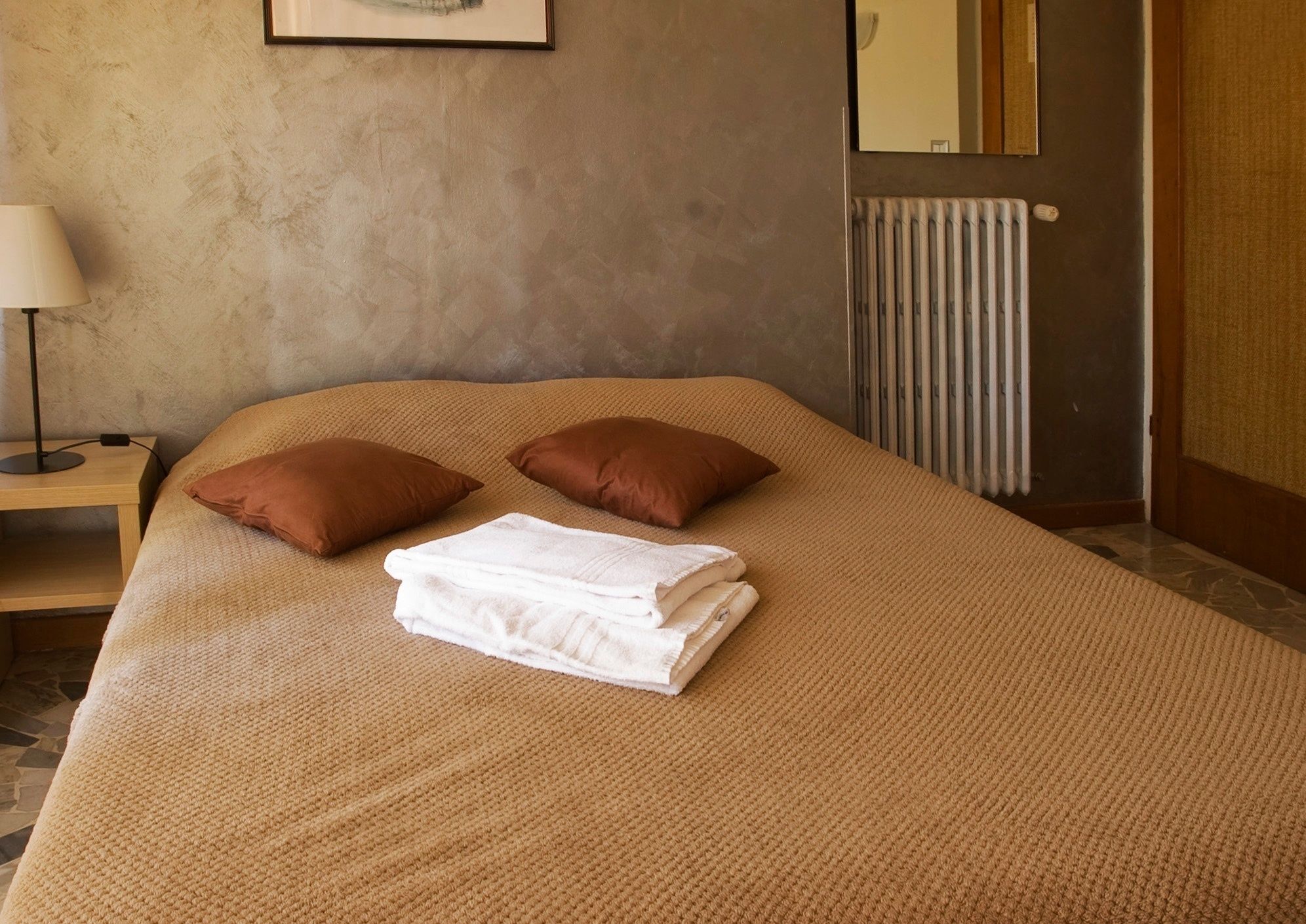 Una cama bien hecha con almohadas marrones y toallas blancas dobladas, una lámpara de noche y un radiador en la pared.