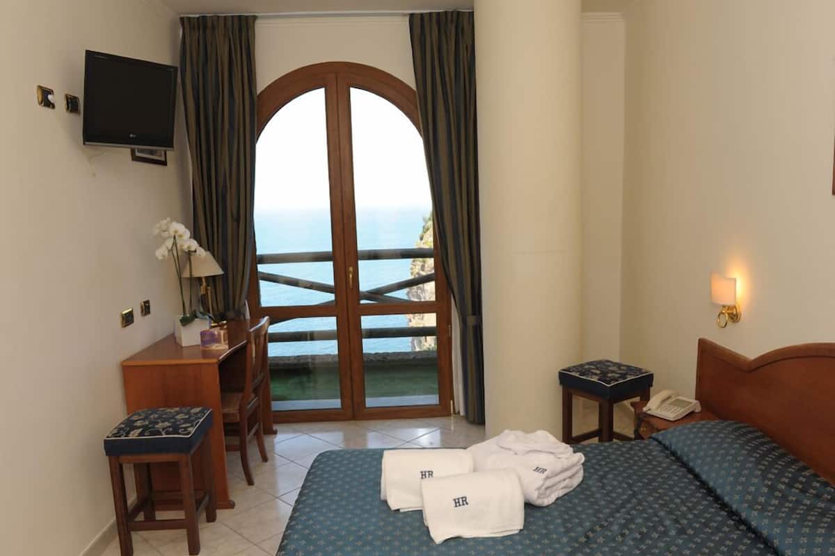 Habitación de hotel con cama, escritorio, silla, televisión, teléfono y vista al mar a través de un ventanal amplio.