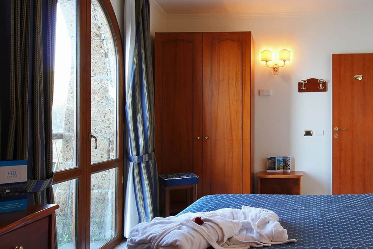 Una habitación de hotel con cama, un armario de madera, una mesita de noche pequeña y una lámpara de pared.